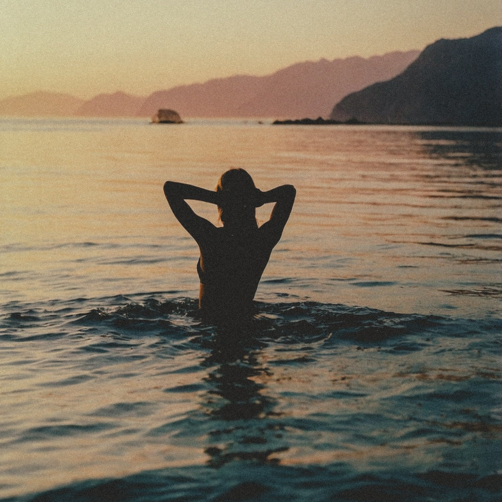 a woman standing in the ocean at sunset looking looking off into the distance